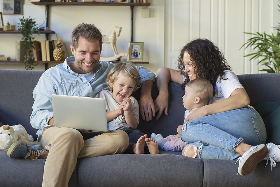 About Our Agency - A Happy Mother, Father and Two Small Children are Smiling and Laughing Together at Home on the Sofa