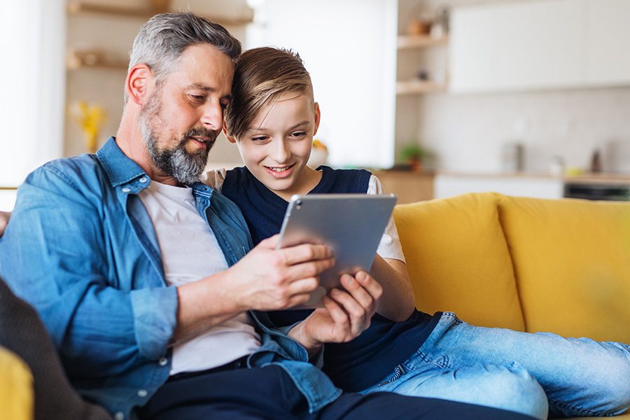 Client Center - Father and Son are Sitting Together on an Yellow Sofa Using a Tablet at Home